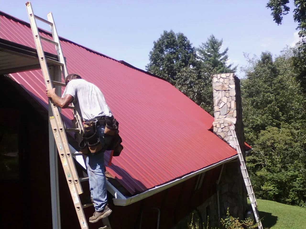 Roofer on a ladder beside a red metal roof and stone chimney — Soffit & Fascia Repair by Roofing Brothers in Beaver Creek